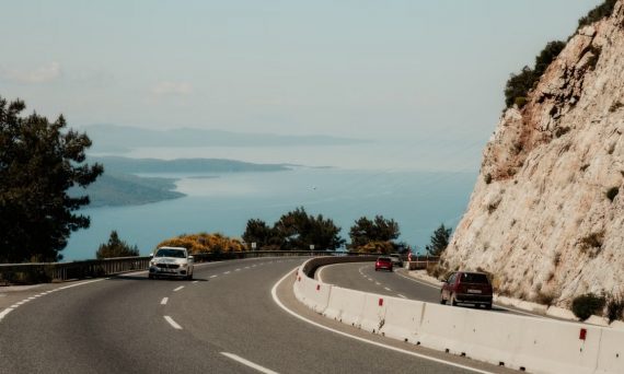 A winding coastal road with cars, overlooking a serene sea and rocky cliffs.